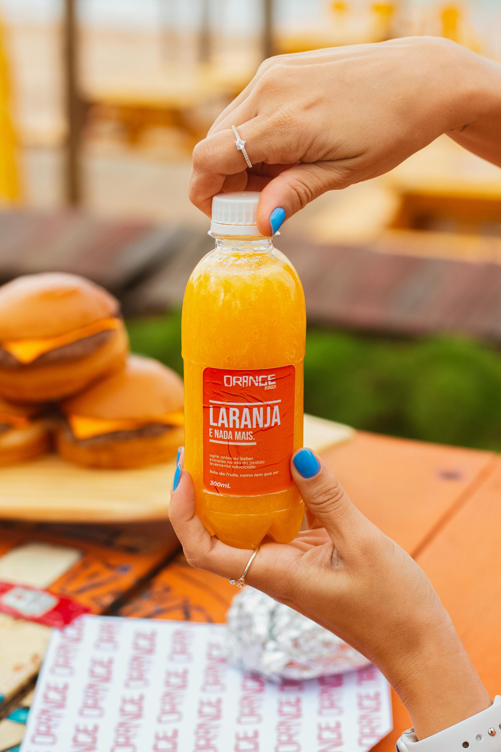 Close-up of hands opening a bottle of refreshing orange juice outdoors with burgers in the background.