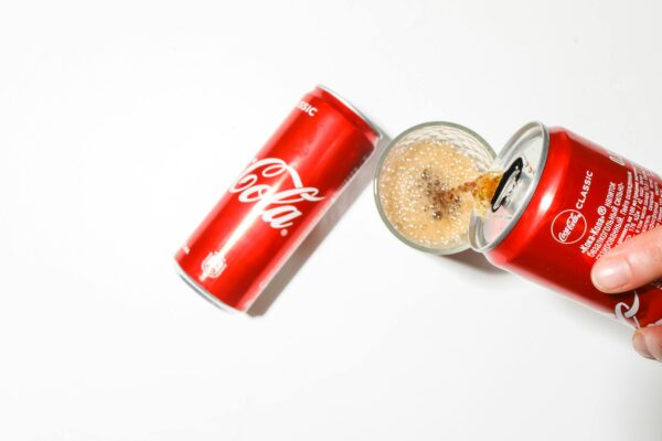 A can of cola being poured into a glass with bubbles forming, captured on a white background.