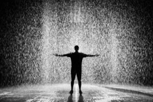 A person joyfully standing in the rain with arms wide open in a dramatic black and white scene.