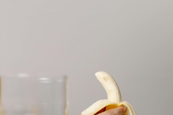 A minimalist photo of a hand holding a peeled banana next to a glass of water on a white surface.
