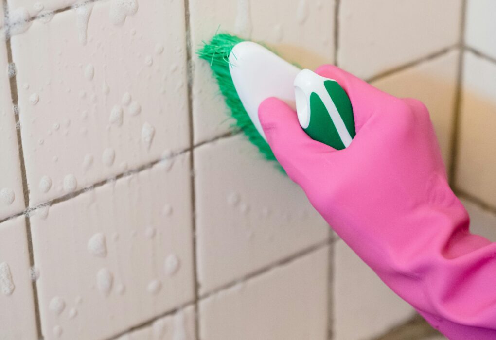 A hand in pink gloves scrubs bathroom tiles with a green brush to remove grime.