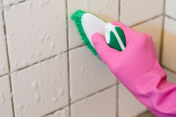 A hand in pink gloves scrubs bathroom tiles with a green brush to remove grime.
