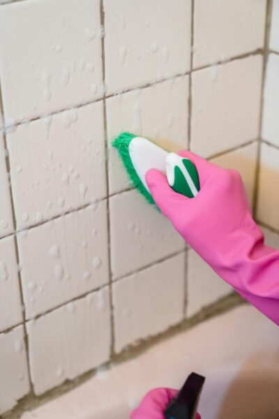 A hand in pink gloves scrubs bathroom tiles with a green brush to remove grime.